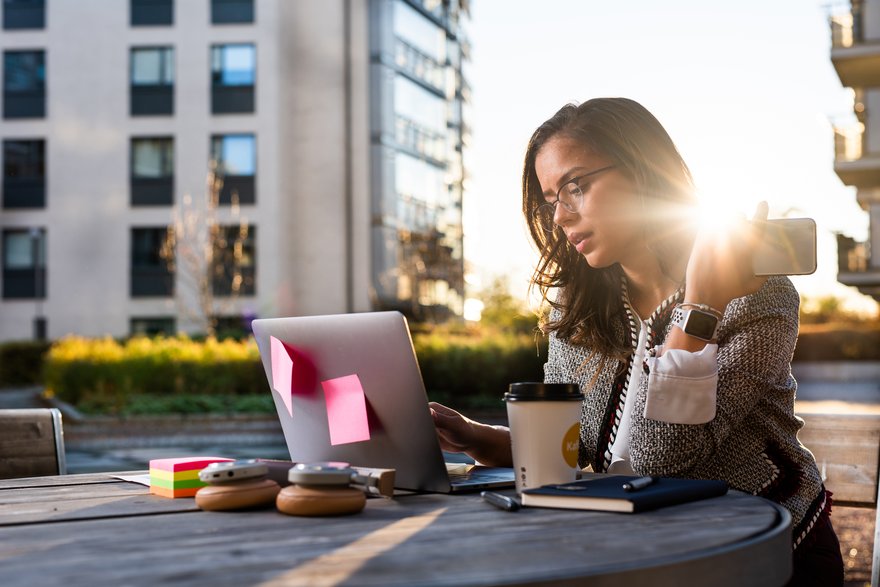Woman working outdoors with laptop, phone, and headphones.