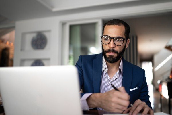 Adult wearing glasses takes notes thoughtfully while sitting at desk with laptop.